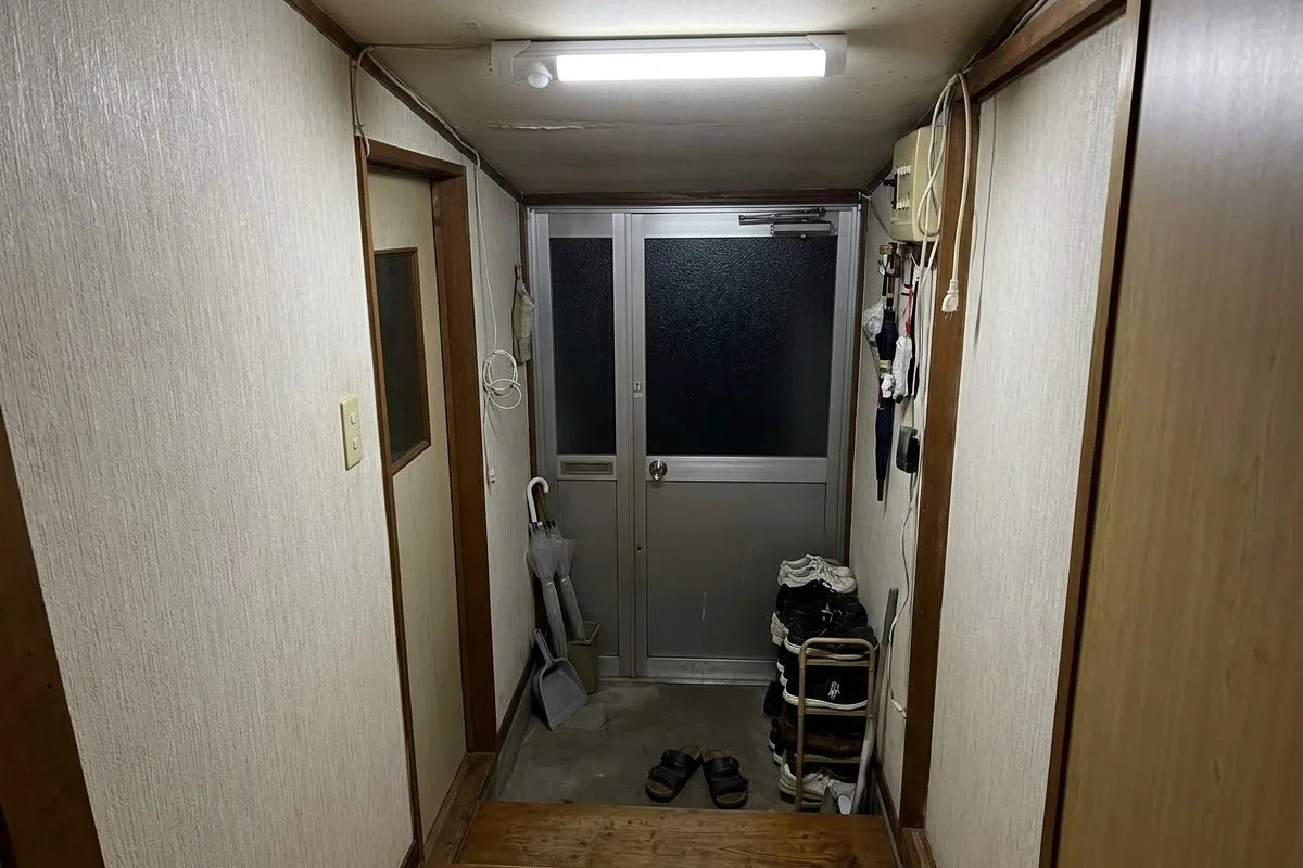 勝手口の内側の土間とアルミの扉。低い天井の直管の器具が1台、昼光色で灯っている